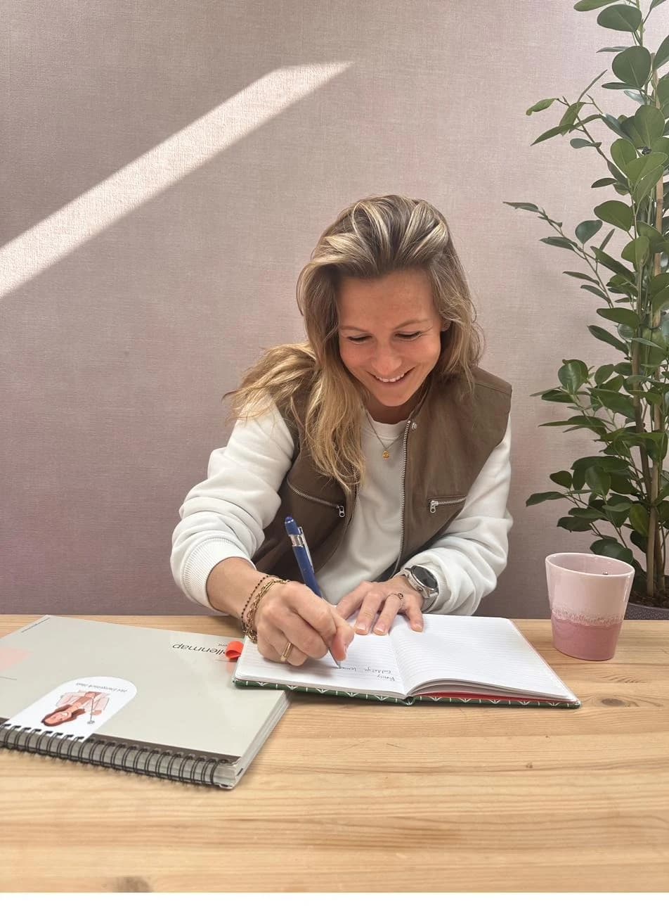 Debbie schrijft aan tafel in haar notitieboek in een ruimte met daglicht